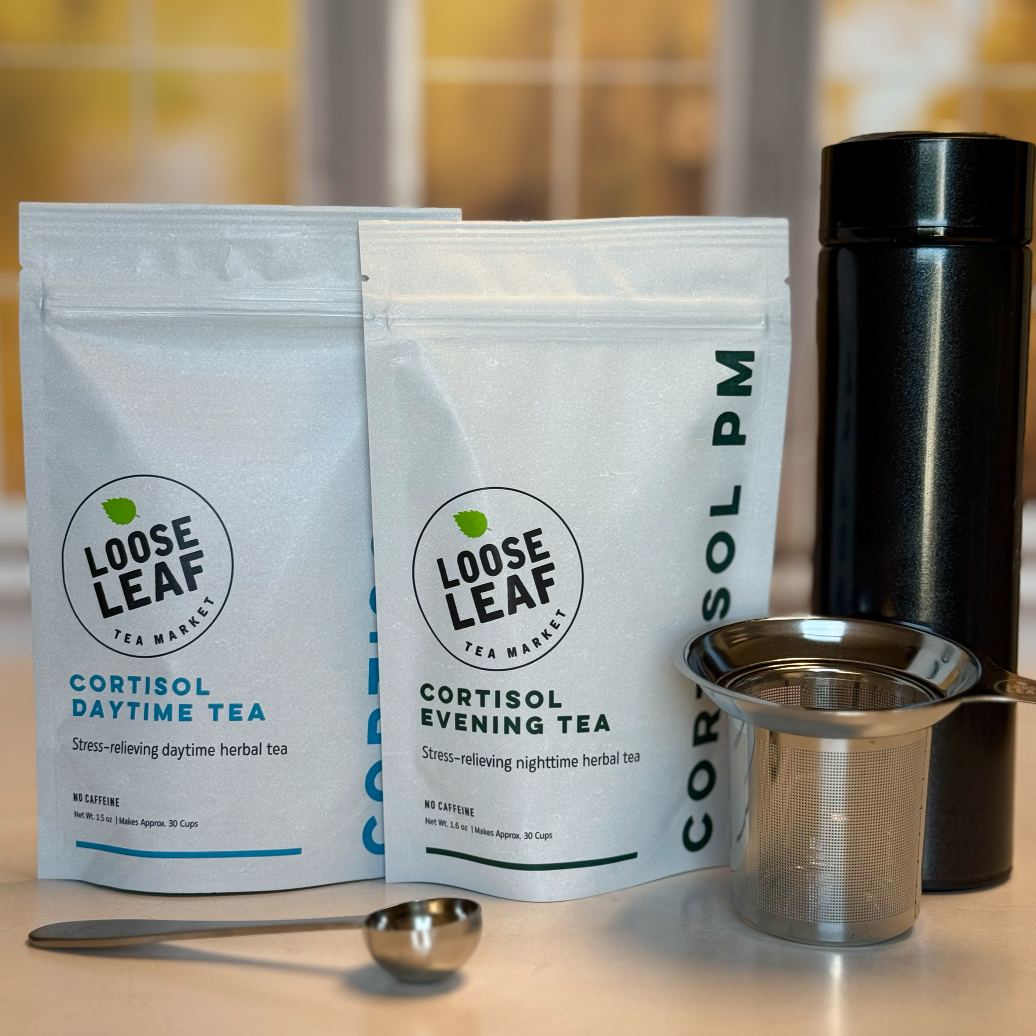 The two teas in the Cortisol Tea Bundle with a stainless steel tea scoop, stainless steel tea infuser, and travel tea tumbler.