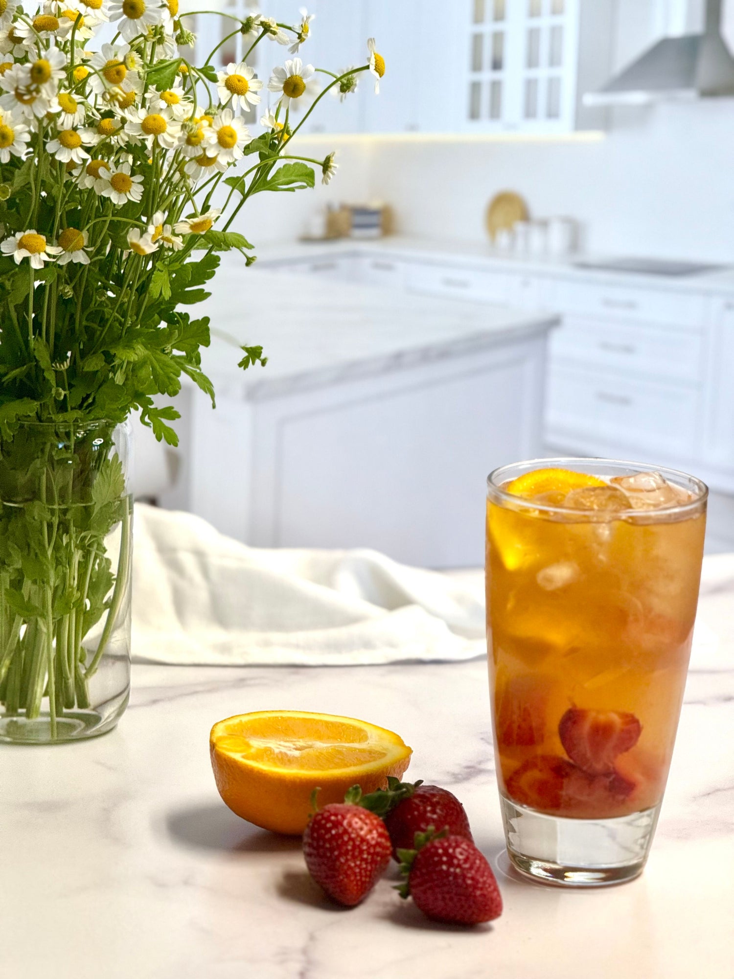Glass of Cortisol Daytime iced tea with strawberries and oranges on a kitchen counter with flowers.