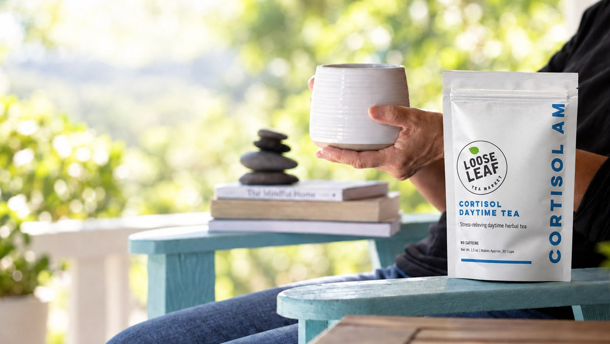 A hand holding a white mug beside a Loose Leaf Tea Market Cortisol Daytime Tea pouch on a porch chair