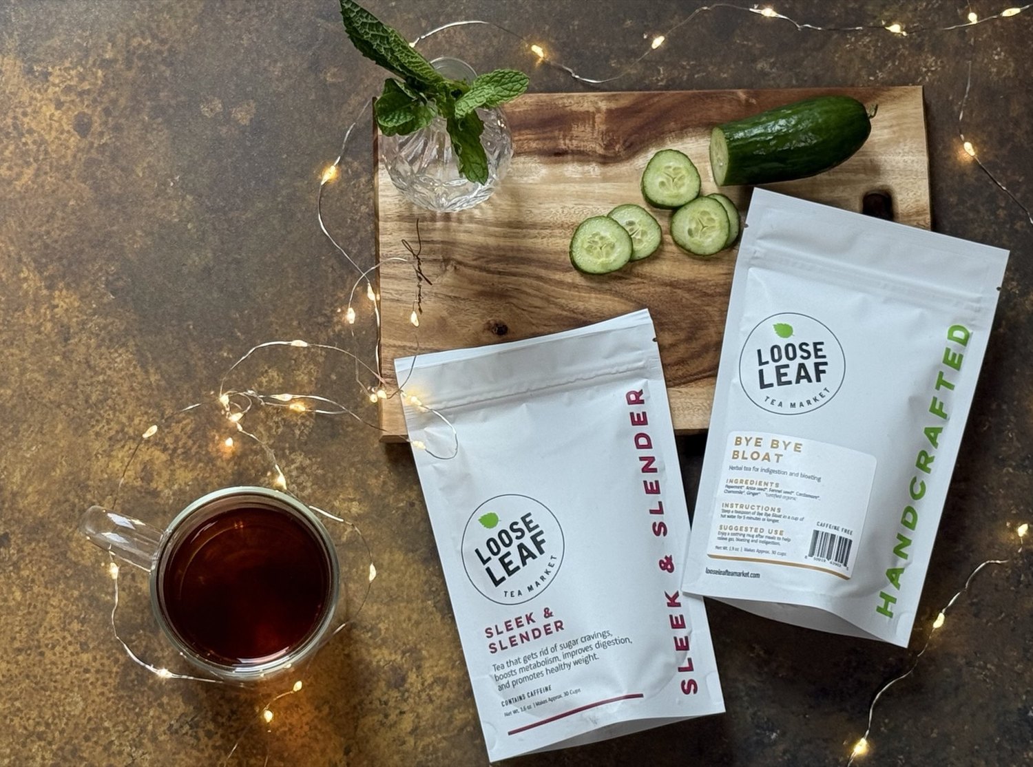 Sleek & Slender and Bye Bye Bloat pouches beside a cup of tea, fresh mint and sliced cucumber